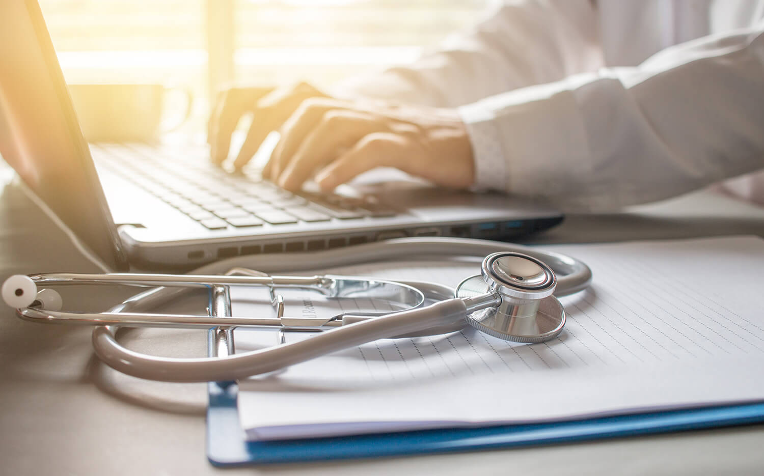 doctors hands typing on laptop; stethescope and notepad on table